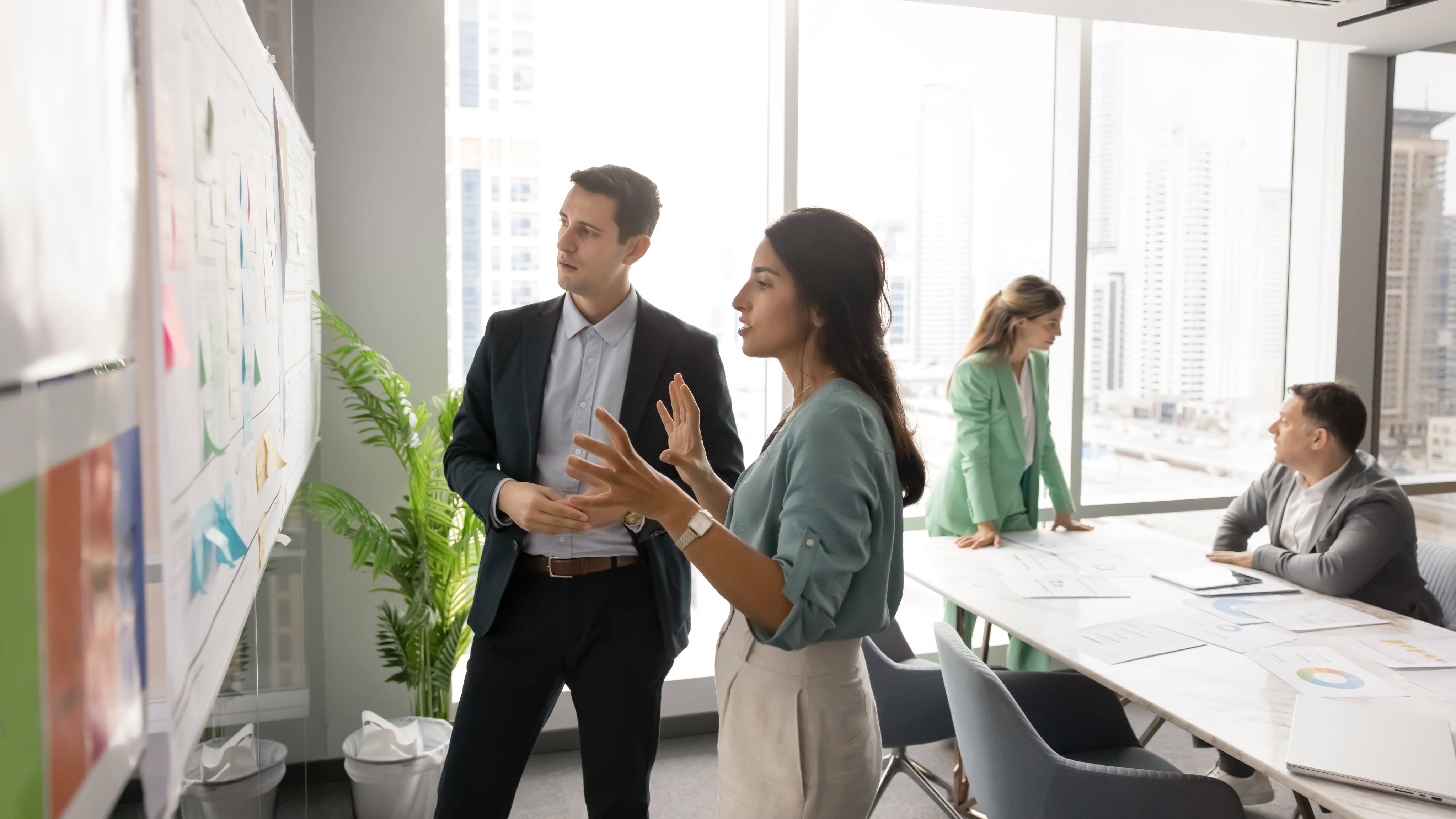 In modern office with city view, woman and man standing near whiteboard filled with sticky notes, female explain or present idea to colleague, reviewing documents, strategizing together in boardroom